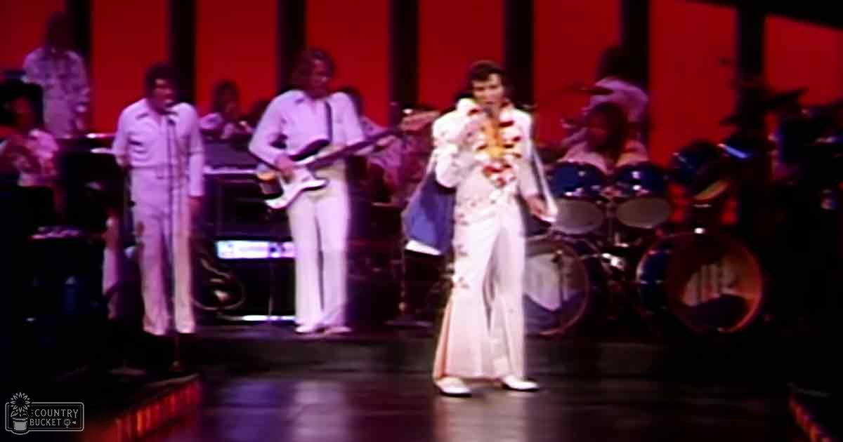 A young adult male performer in a white jumpsuit with gold floral patterns and high collar is on stage at an indoor venue, possibly a theater or concert hall, accompanied by musicians dressed in light-colored suits from the mid-20th century.