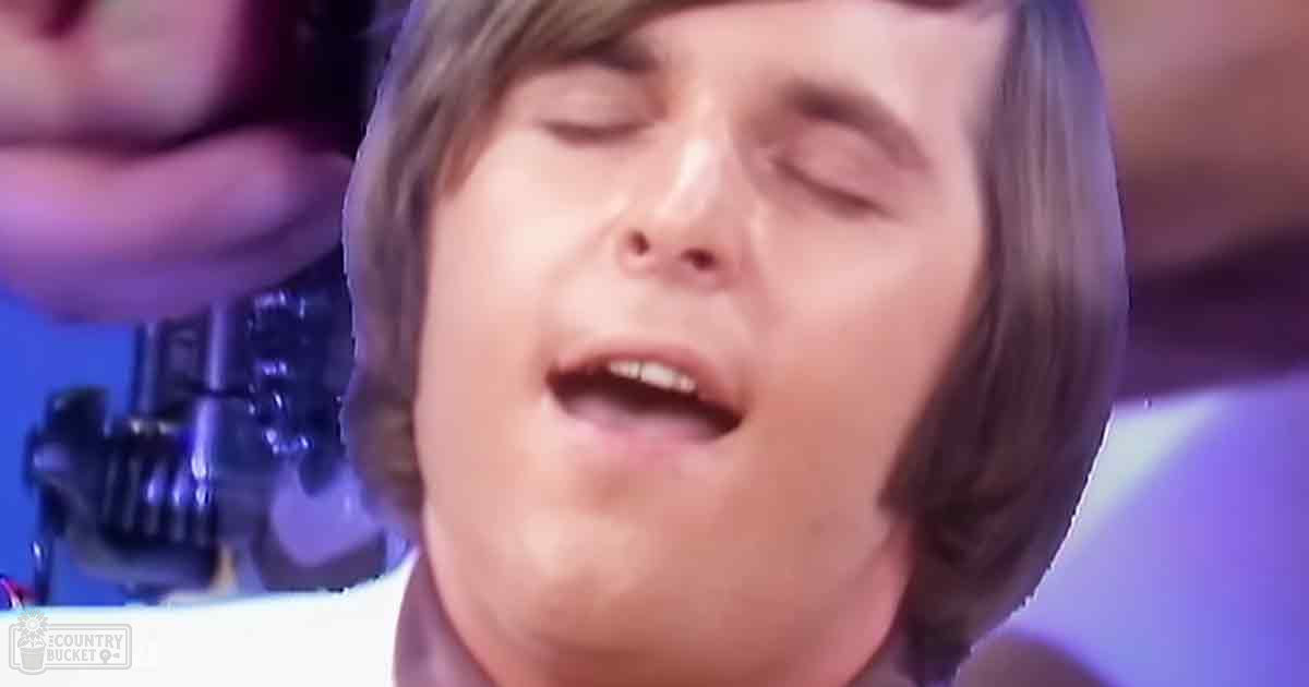 A young adult male singer in a white collared shirt, performing on stage with purple lighting and equipment visible behind him.