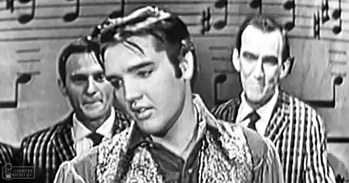 A young adult male in patterned shirt with scarf or bandana around neck stands in an older film set, possibly a school or institutional environment; two adults are partially visible behind him, wearing formal attire from earlier eras.