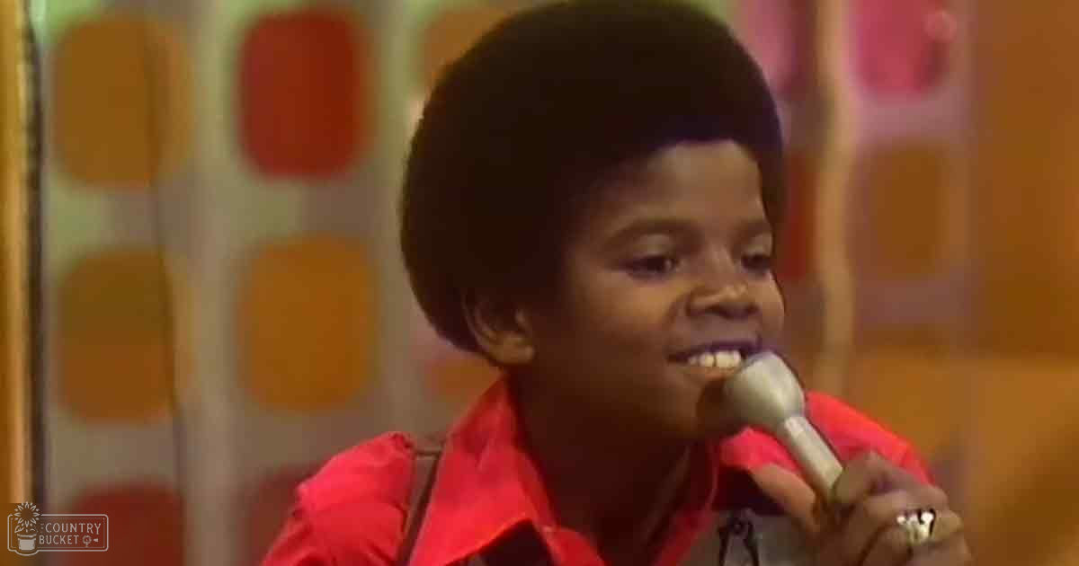 A young child wearing a bright red button-up shirt or dress sings into a microphone on stage with colorful geometric patterns behind them.