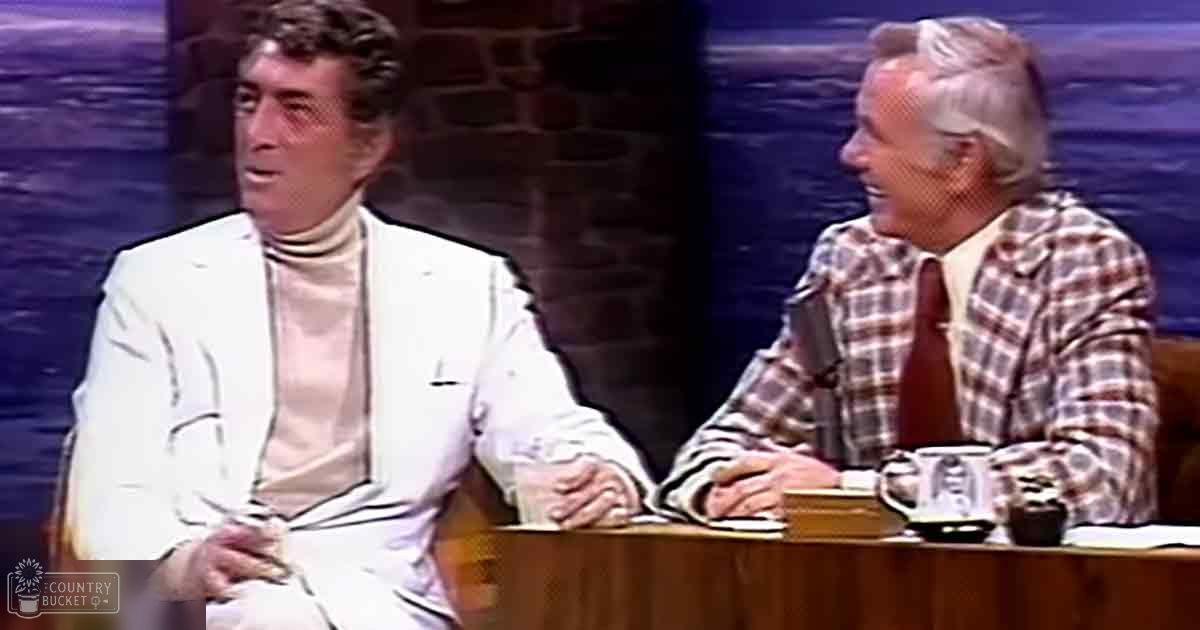 Two individuals seated at a table on a talk show set against a dark blue backdrop with stone column structures, suggesting an indoor studio environment.
