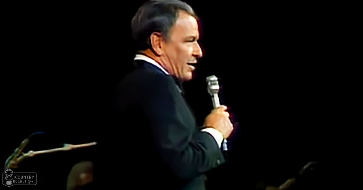 A middle-aged male singer in a dark mid-20th-century-style suit jacket and tie, singing into a microphone on stage under focused stage lighting.