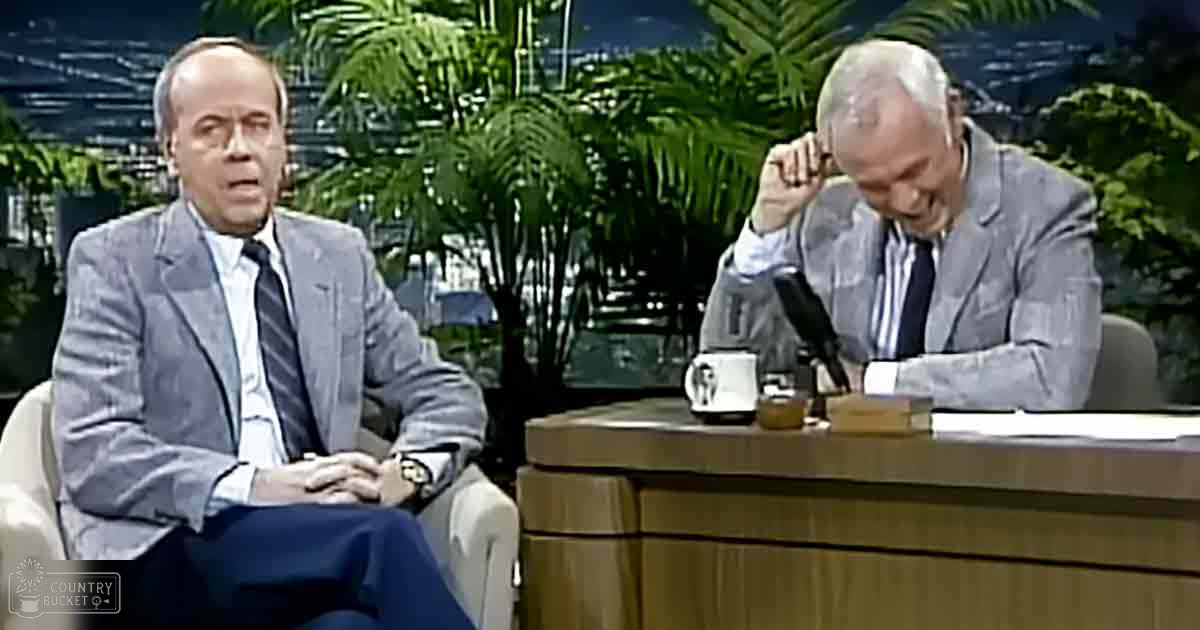 Adult male talk show host interviewing older adult guest in a mid-20th century indoor studio with wooden desk, potted plants, and cityscape visible through windows.