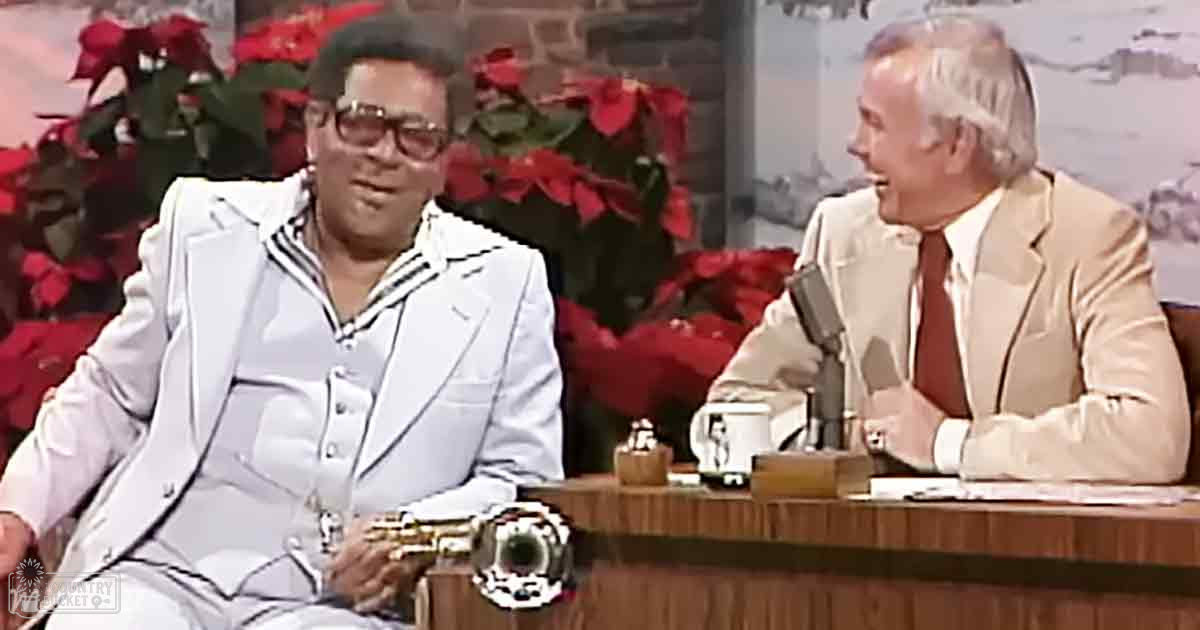 Two adult male hosts on a talk show set, one in a white suit with black stripes and holding an instrument, another in a beige suit with a red tie. The indoor studio features wooden furniture, poinsettias, brick wall, and artificial lighting.