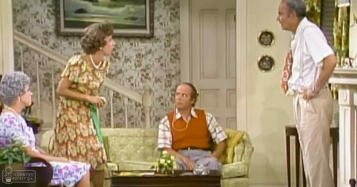 A woman in her adult years stands left, wearing a floral dress with pearl necklace; another woman sits right, dressed casually. A man is seated on the green patterned sofa, adorned with an orange vest and gold chain.