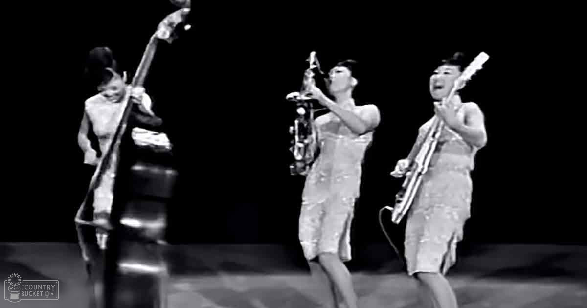 Musical performance scene on a stage, young adult females in sleeveless dresses holding string and wind instruments against dark curtains at an evening venue.