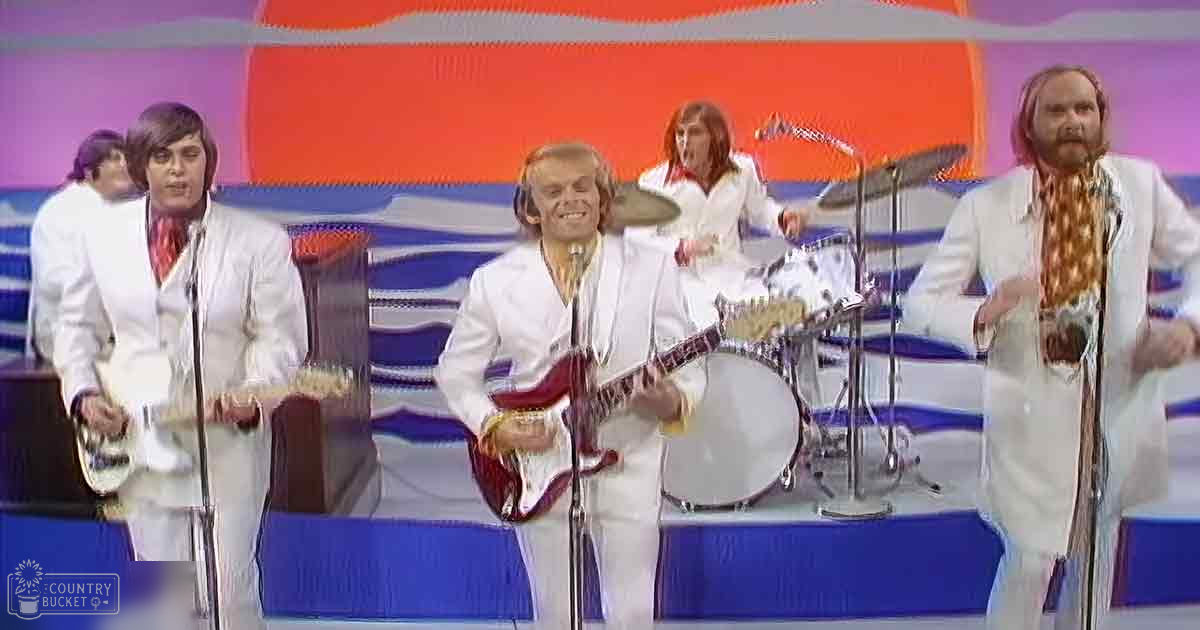 A musical performance scene from the 1970s with performers on stage in formal white suits and colorful scarves, set against a vibrant abstract backdrop.