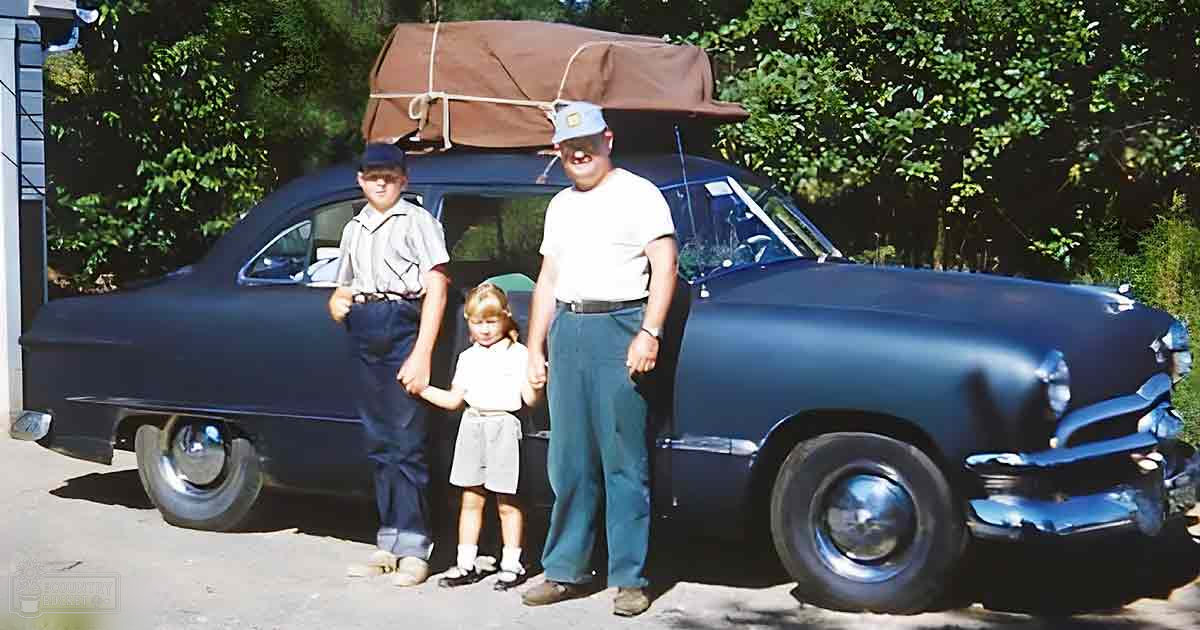A middle-aged adult male wearing a white T-shirt and dark blue trousers stands with children near an old classic car on a sunny day in a residential suburban setting.