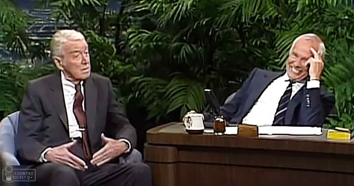 Two older adult males in conversation within an indoor talk show set with lush green foliage, suggesting a tropical theme and well-lit studio environment.