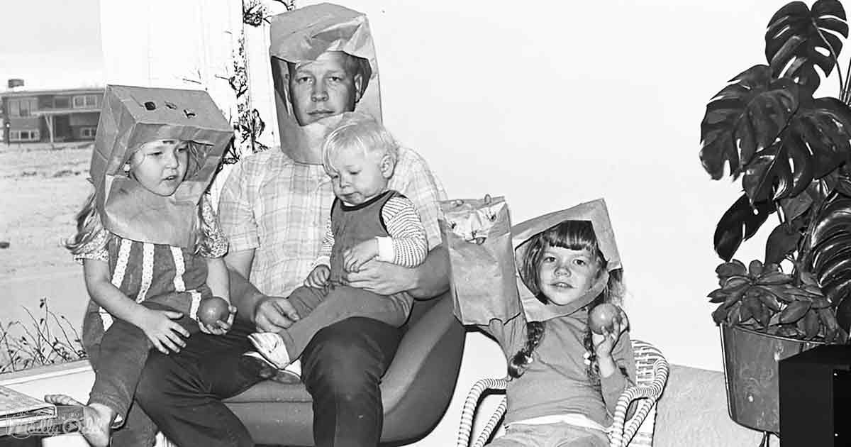 A family scene indoors with adults and children wearing paper bags over their heads, seated on a wicker chair in front of a wall and part of an outdoor view through a window or door.