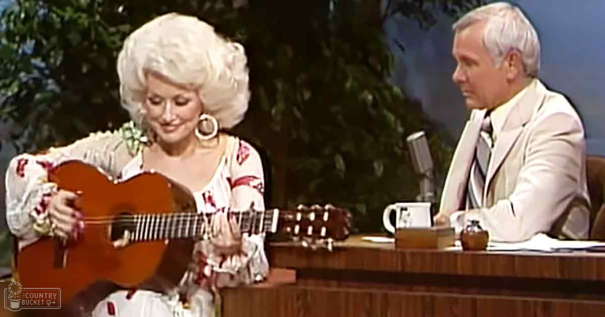 A woman playing guitar in a talk show set with an older adult male host, surrounded by indoor decor that mimics an outdoor environment.