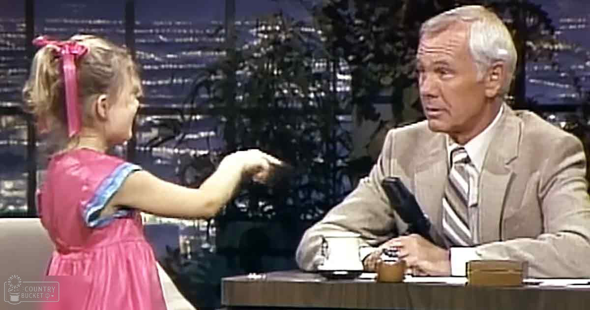 A young girl wearing a pink dress with blue trim and a red ribbon interacts with an older adult male in a light-colored suit jacket at a desk, with city lights visible in the background. The setting appears to be an urban talk show studio recorded at night.