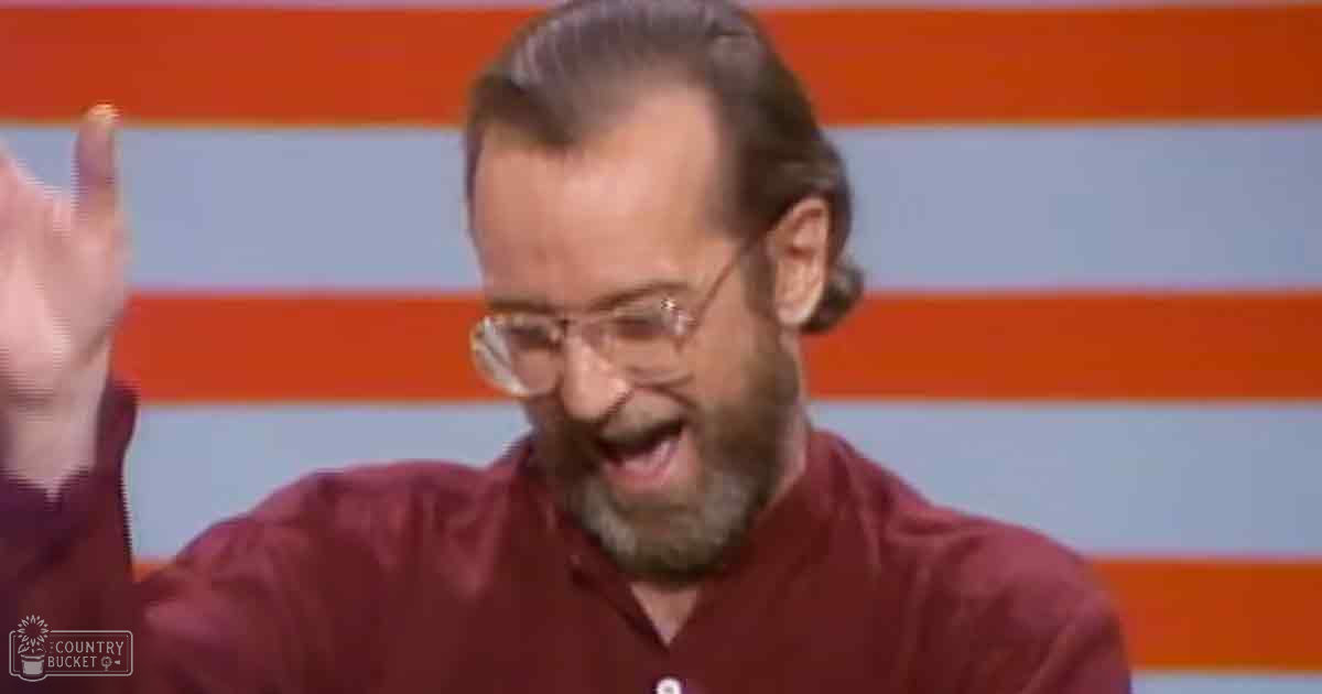 A middle-aged adult male wearing a maroon button-up shirt in an indoor setting with orange and white striped background resembling a stage or set design.