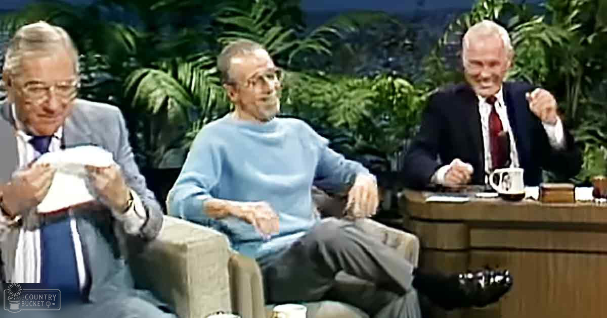 Three older adult males seated in a talk show setting with lush green foliage and bright indoor lighting, discussing or presenting.