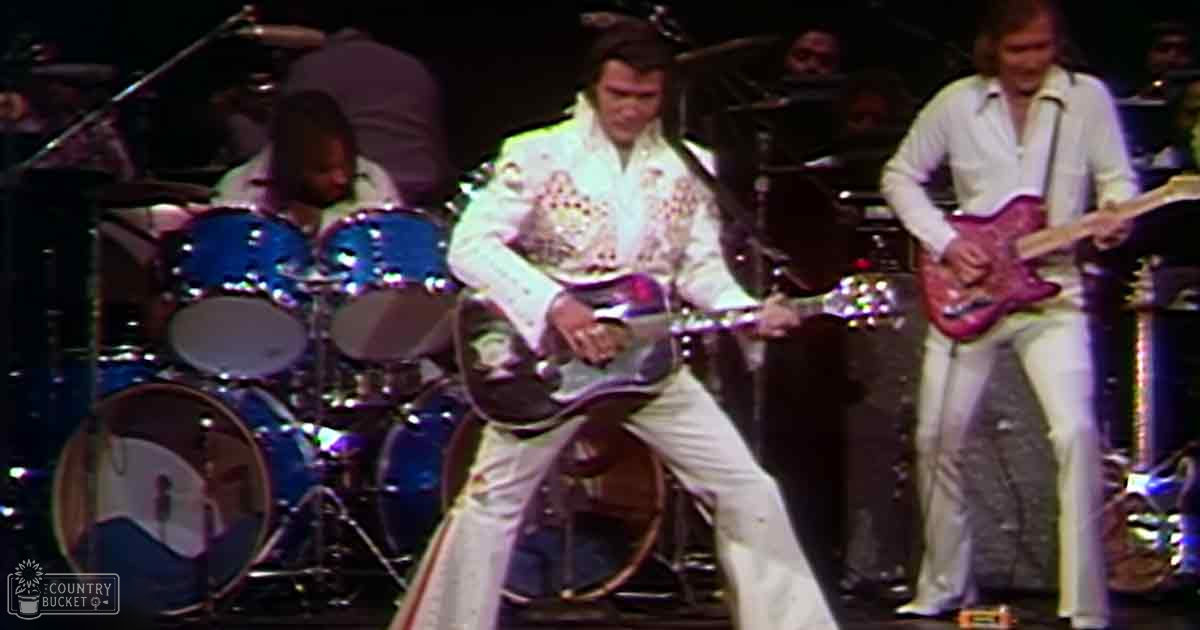 Adult male performers wearing white outfits with decorative patterns, playing electric guitars on a stage with a drum set in the background during a live music performance.