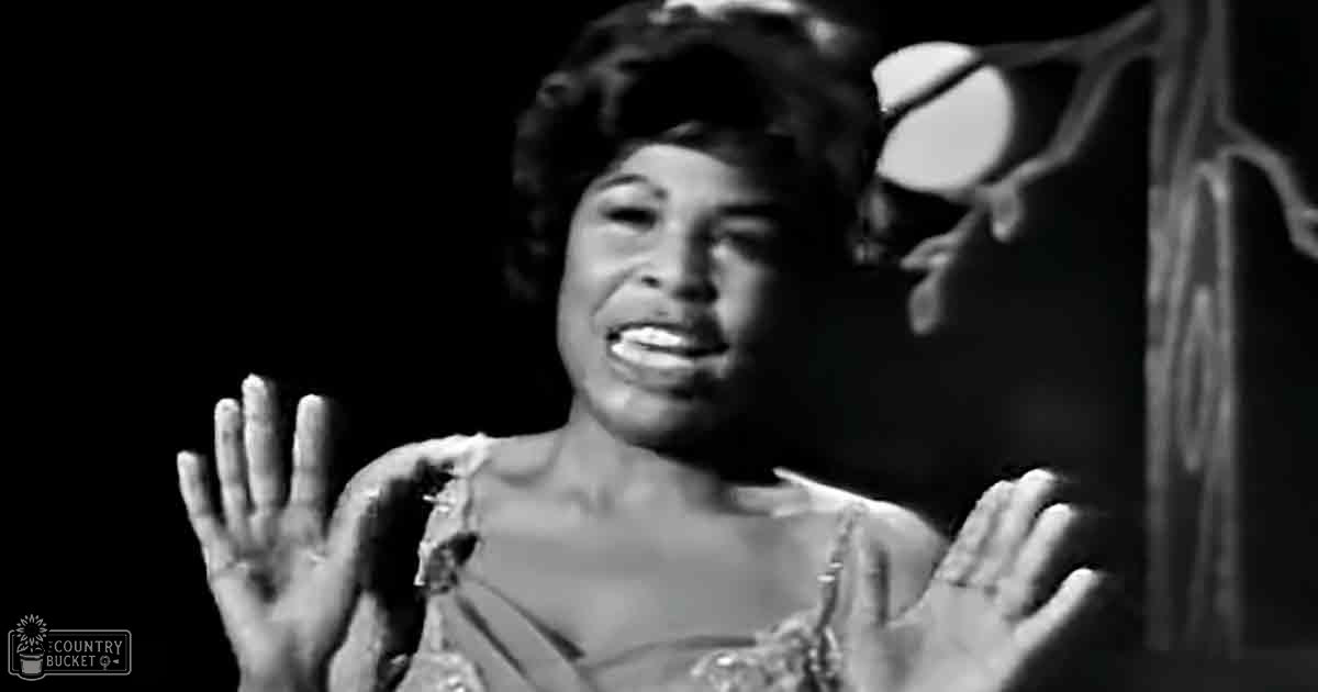 An adult female performer in a mid-20th century sleeveless, low-necked garment with decorative elements on shoulders, wearing bracelet, indoors under dramatic lighting.