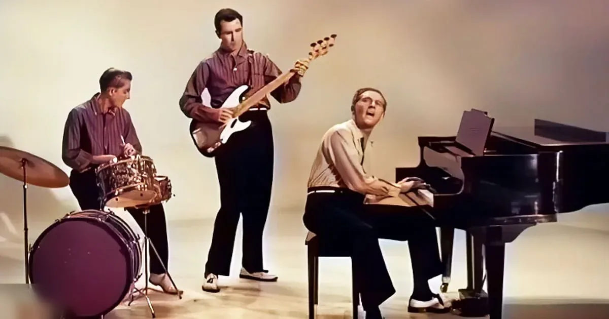 Musical performance with a young adult male drummer, an adult male bass guitarist, and a middle-aged to older male pianist/singer in a studio setting.