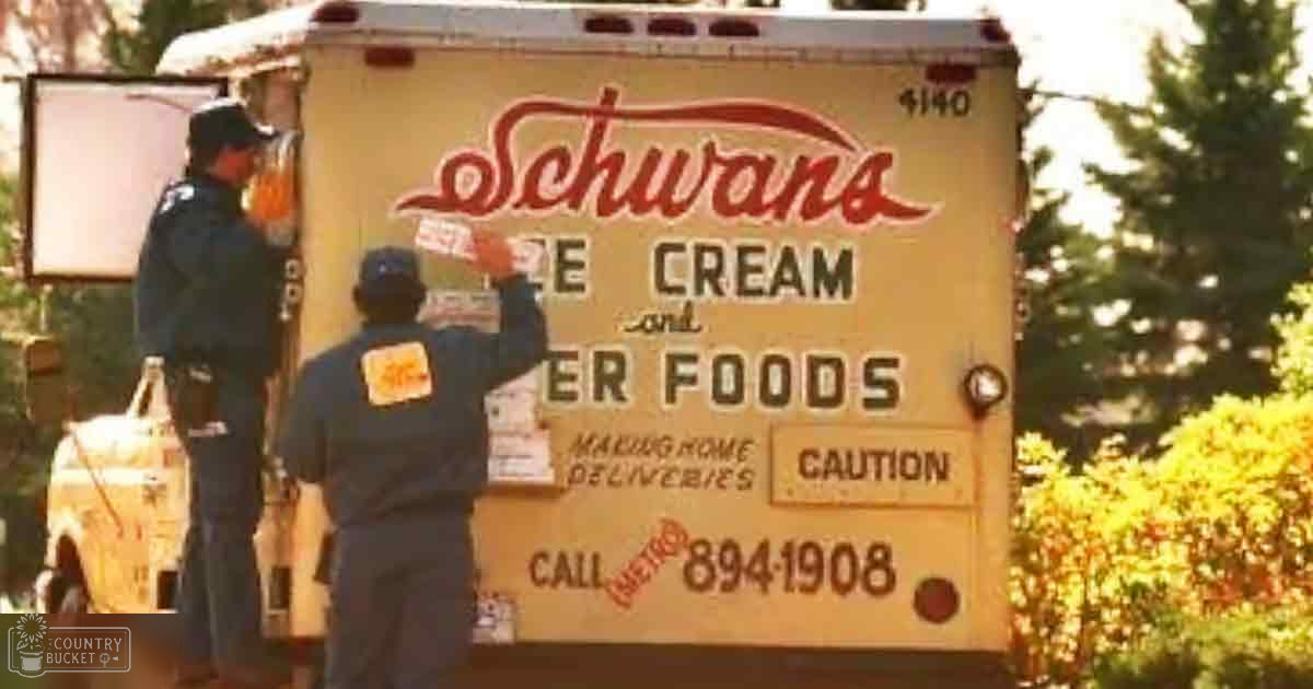 Two adult males in dark blue uniforms unloading a Schwans Ice Cream and Other Foods delivery truck in an outdoor residential area with trees and bushes, late afternoon light.