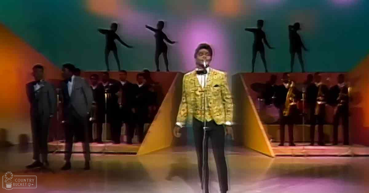 Adult male singer wearing a shiny gold jacket with intricate patterns, dark pants, and a bow tie on stage during an evening performance.
