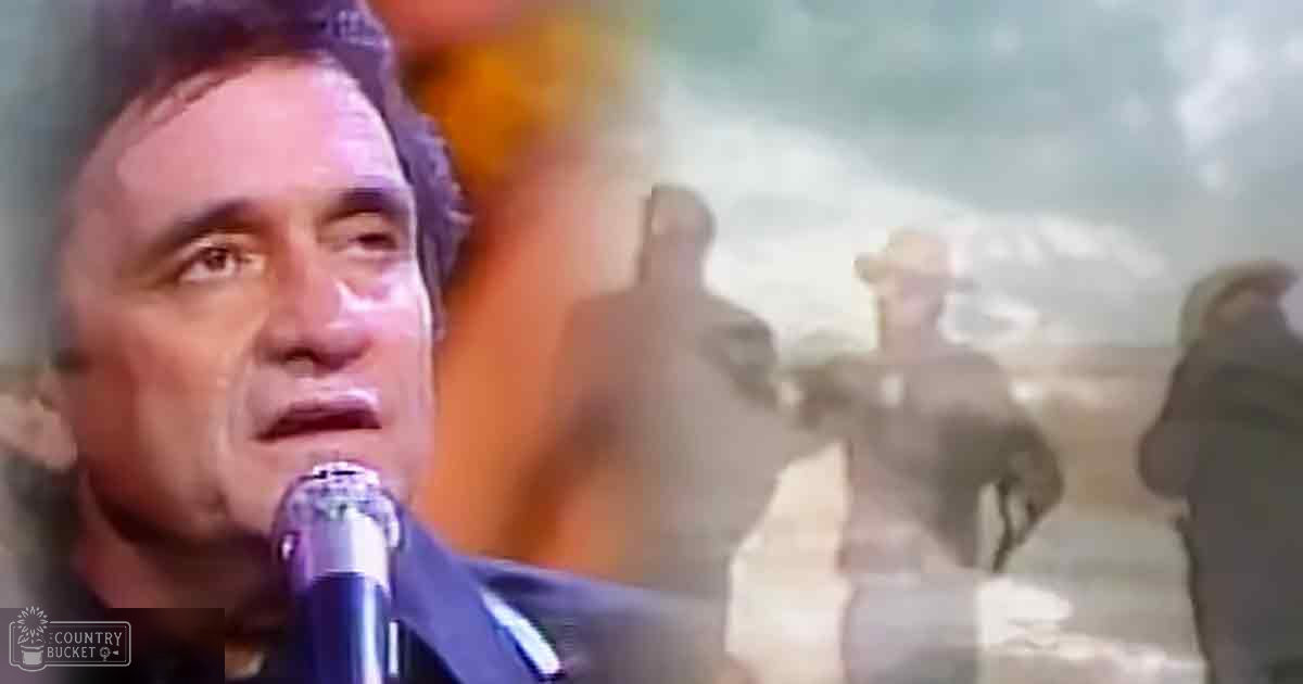 An adult male singer wearing a dark suit jacket and shirt sings into a microphone against an outdoor backdrop with silhouettes of people, suggesting a concert setting.