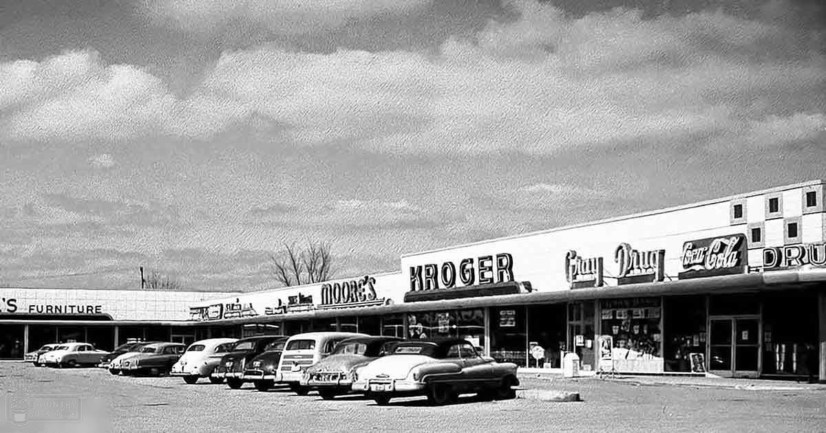 An older suburban shopping center with vintage cars parked in front of stores on a cloudy day.