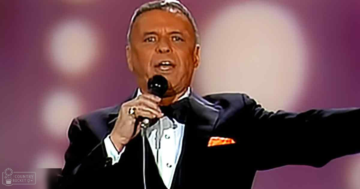 A middle-aged male performer in a well-fitted black tuxedo with white lapels, holding a microphone on an indoor stage at night.