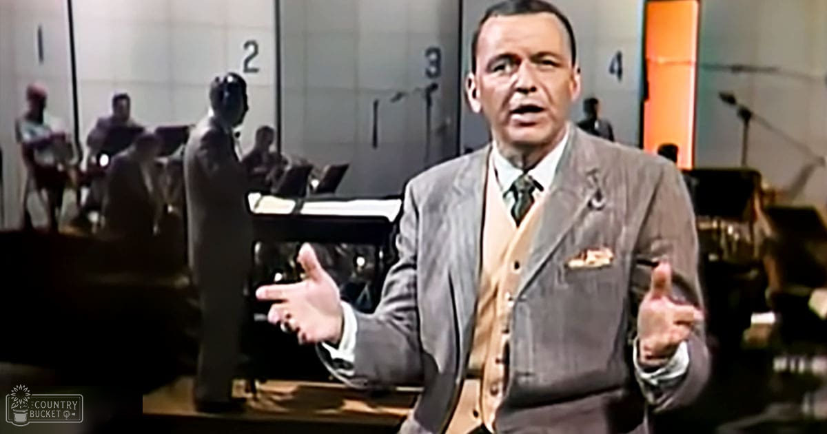 A middle-aged adult male wearing a gray suit with a beige vest and tie, white shirt, and pocket square gestures in an indoor studio or stage setup for a performance or recording session. Bright artificial lighting illuminates the scene.