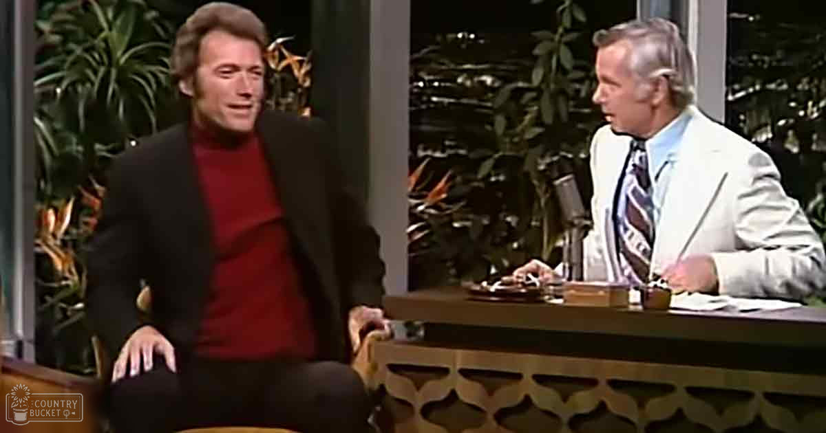 A young adult male guest wearing a red turtleneck sweater under a dark blazer, seated on the left in a mid-20th-century style talk show studio with an older man in a light-colored suit and patterned tie. The setting includes a wooden desk with microphones, plants, and nighttime cityscape views.