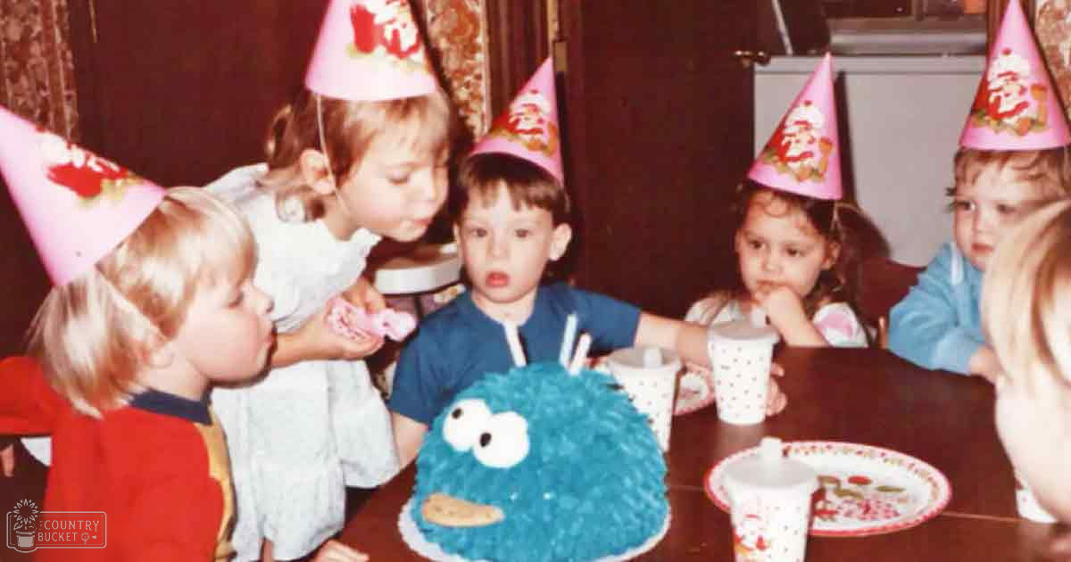 Children gathered around a table at an indoor birthday party with curtains, wearing festive outfits and party hats.