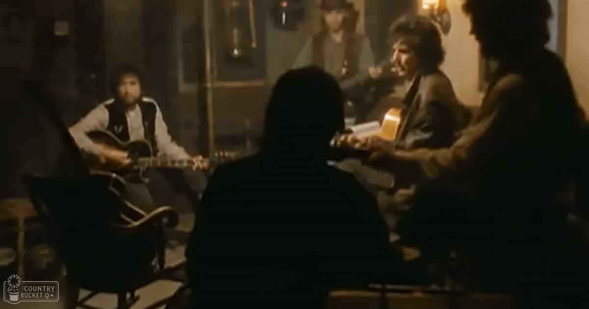 Musical performance indoors with dim lighting, late evening or night ambiance; adult male singer in dark striped jacket, young adult playing acoustic guitar, middle-aged female musician in dark outfit. Wall lanterns and wooden structure suggest old-fashioned bar or pub setting.