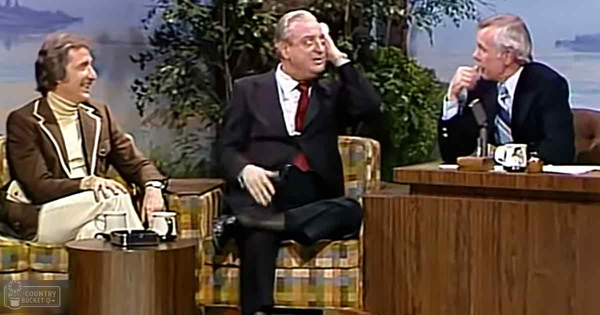 A talk show set with three adult males in formal attire, against a backdrop of foliage and water, suggesting an outdoor setting used as a visual background.