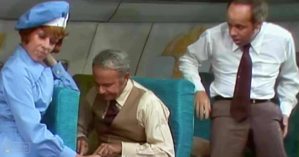 An older adult male seated indoors, wearing a light-colored shirt under a brown vest with a dark tie. Two other individuals in professional uniforms stand nearby, suggesting an airport or train station setting.