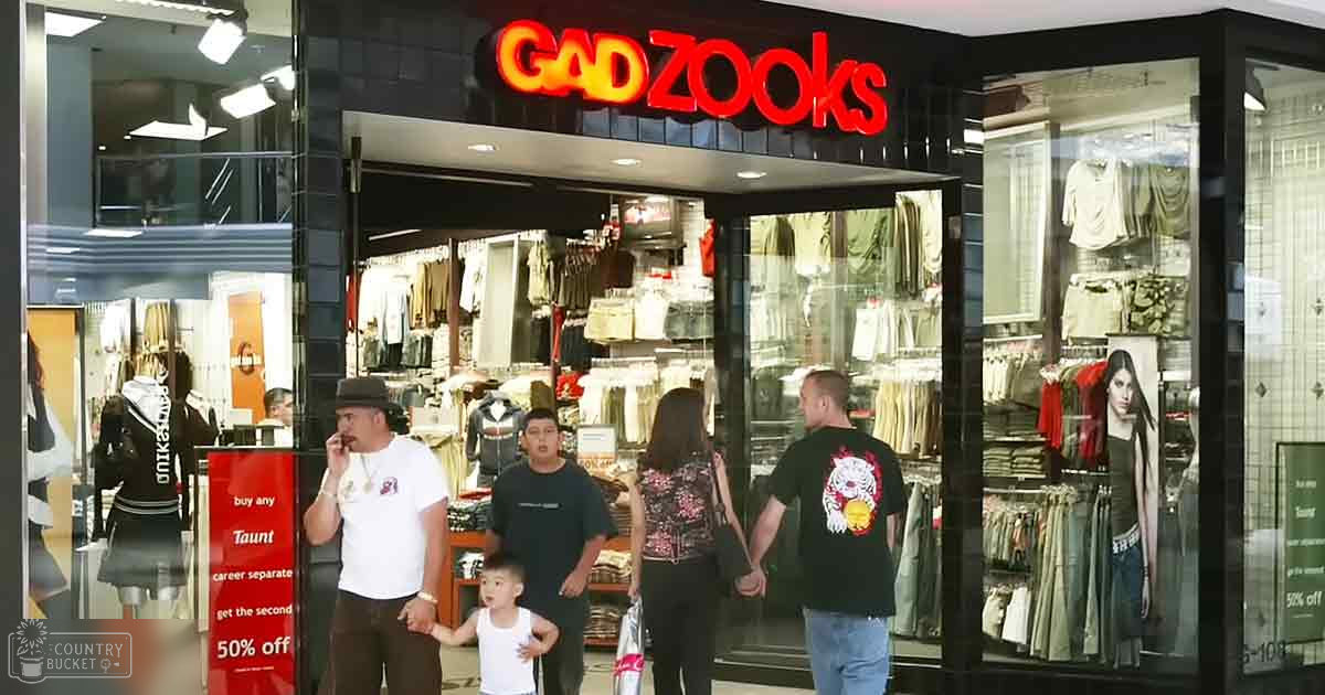 Adult male middle-aged wearing a white t-shirt with red text, dark pants, and a fedora hat stands in front of Gadzooks retail store. Child in white tank top and dark pants walks nearby.