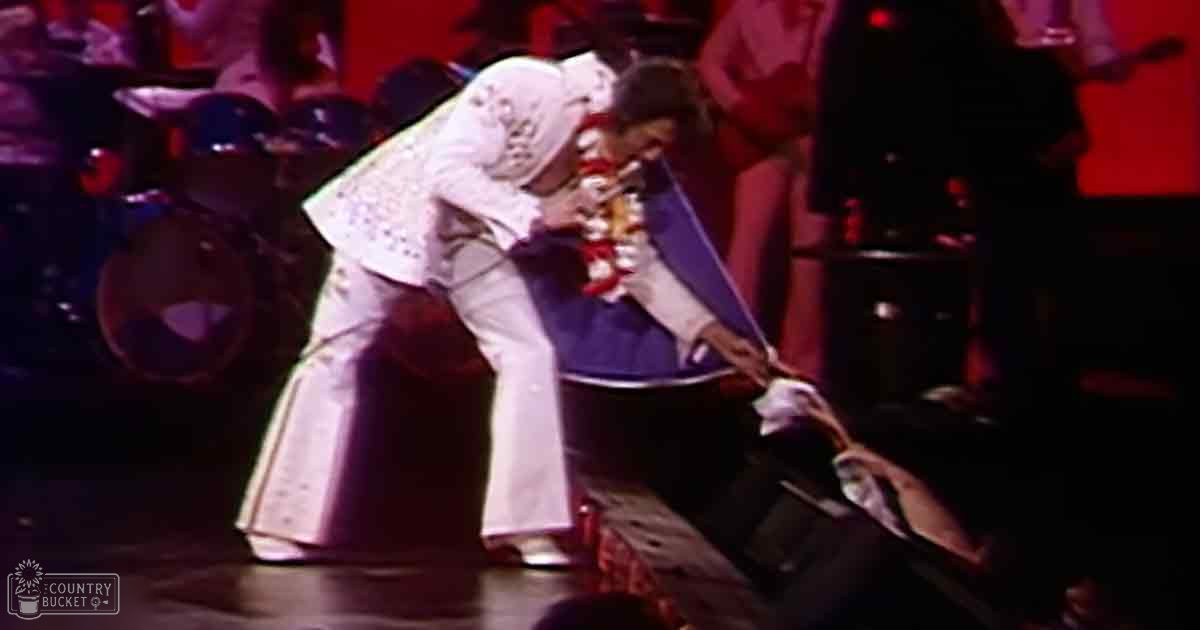 An adult male performer in a white sequined jumpsuit with gloves and shoes, likely from an Elvis tribute show on stage with dimly lit background.