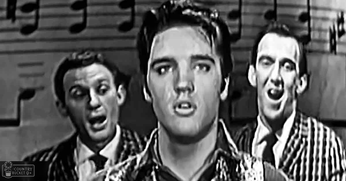 A young adult male singer with short dark hair, wearing striped shirts, stands in front of sheet music on a stage or rehearsal space under even and bright lighting.