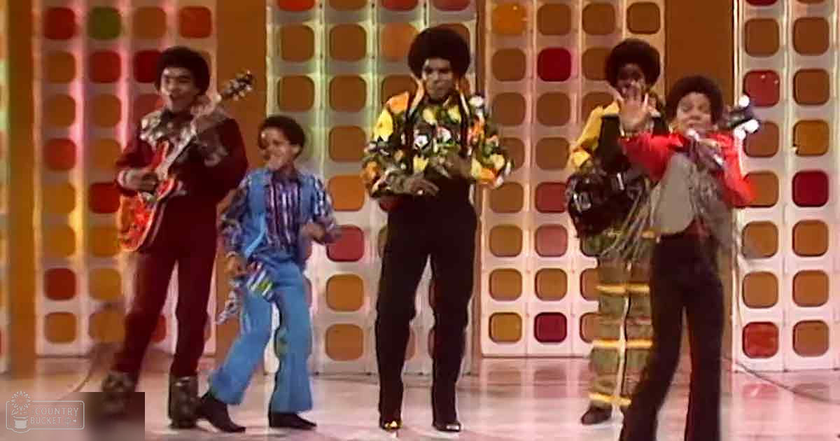 A musical performance group in an indoor studio setting with vibrant geometric backdrop, likely from the 1970s.