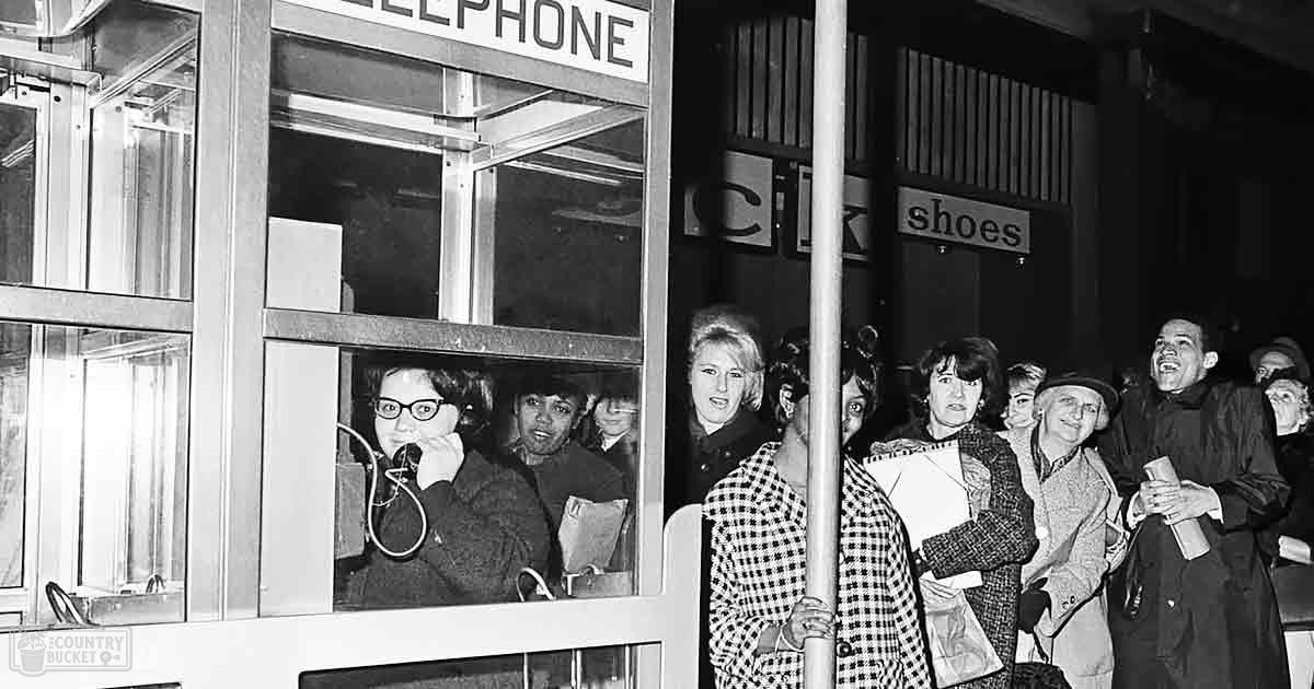 A group of people gathered around a classic-style telephone booth in an urban environment during the evening.