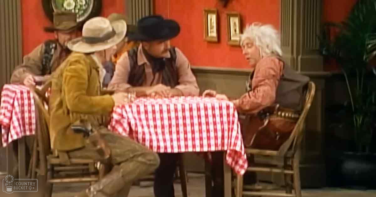 A Western-themed bar or saloon interior with wooden furniture, red-and-white checkered tables, framed pictures on a red wall, and green indoor plants. The setting appears to be indoors during the evening.