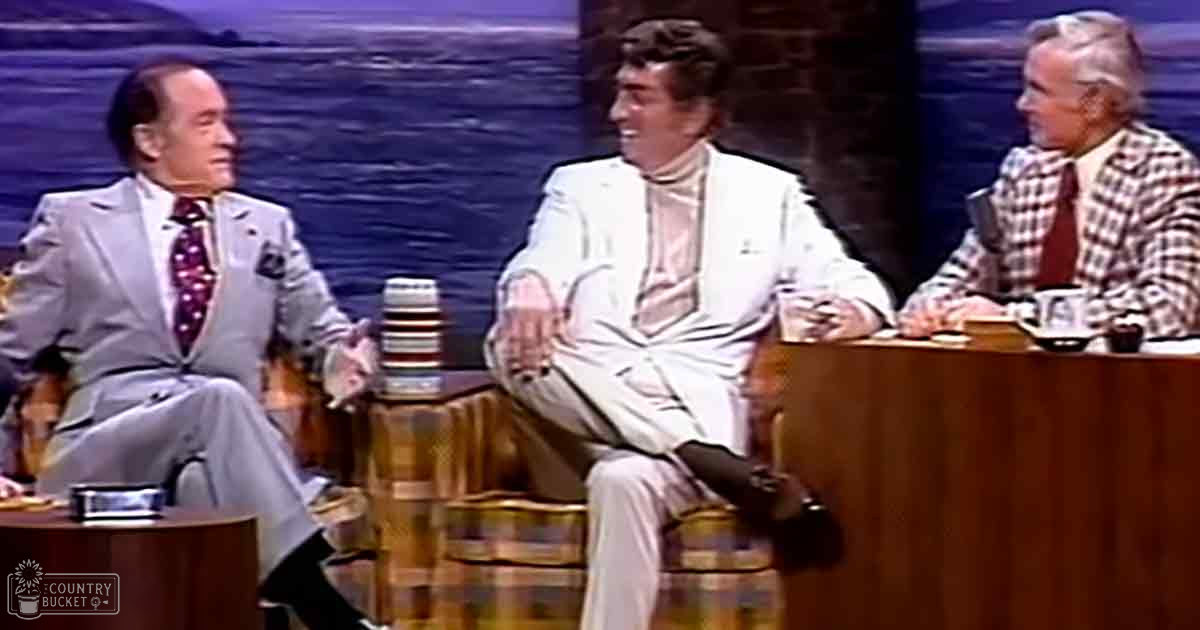A talk show set from the 1970s with three adult male hosts in formal attire seated at a table, against an indoor scenic backdrop suggesting water or a lake.