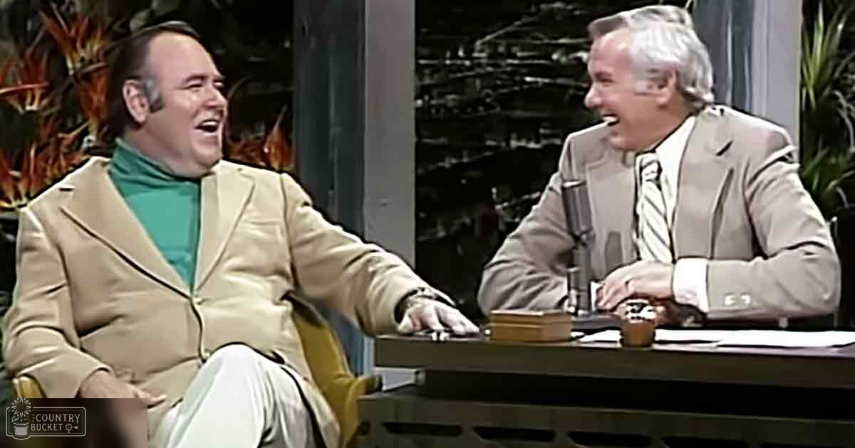 An adult male in a beige blazer and teal shirt with suspenders, seated next to another older adult male in a light-colored suit, in an indoor studio setting with plants visible through large windows.