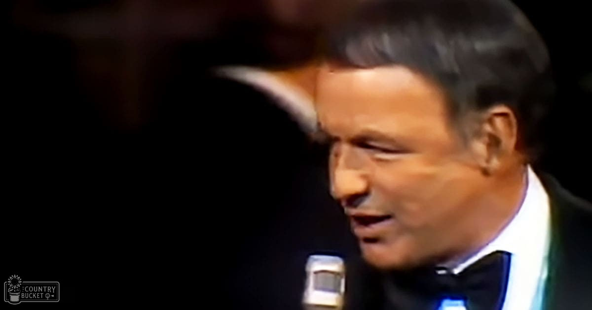 A middle-aged male singer wearing a black tuxedo with white shirt and bow tie, performing on stage indoors under dim lighting.