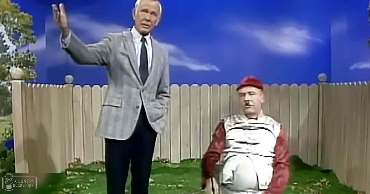 Adult male wearing a gray suit jacket, white shirt, dark tie, black pants standing outdoors with an older adult male seated nearby on grassy terrain.