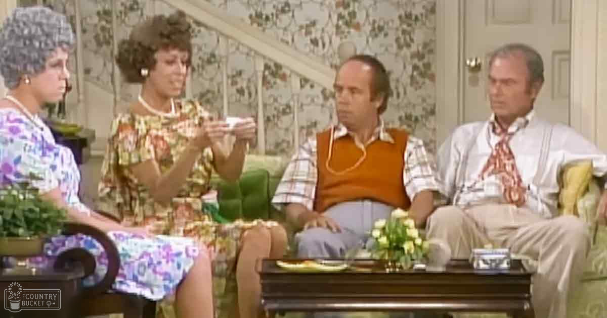Adult female wearing floral dress and pearl necklace in a living room scene with other characters, furniture, and floral wallpaper.