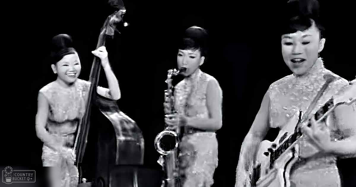 A musical performance scene with young adult females playing a double bass, saxophone, and guitar on stage at night.