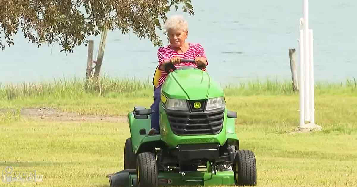 An older adult woman riding a lawn mower near water with greenery and trees in the background, wearing a red and white striped shirt.
