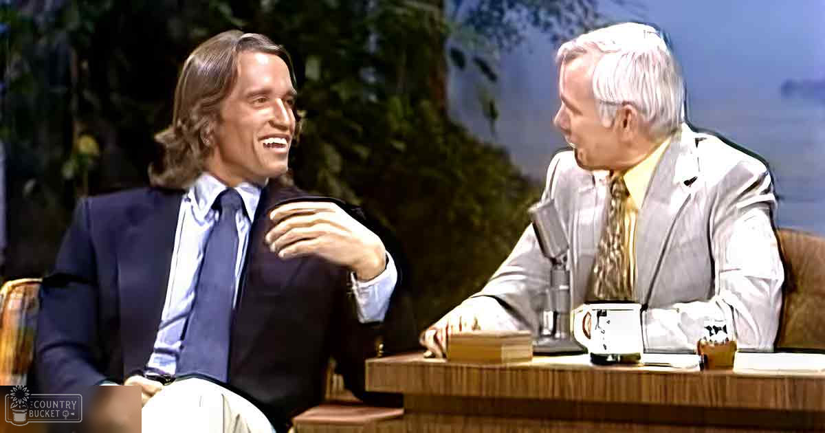 Young adult male in a dark suit jacket over a light blue dress shirt and tie on a talk show set with older adult male in similar attire, greenery backdrop suggesting outdoor setting.