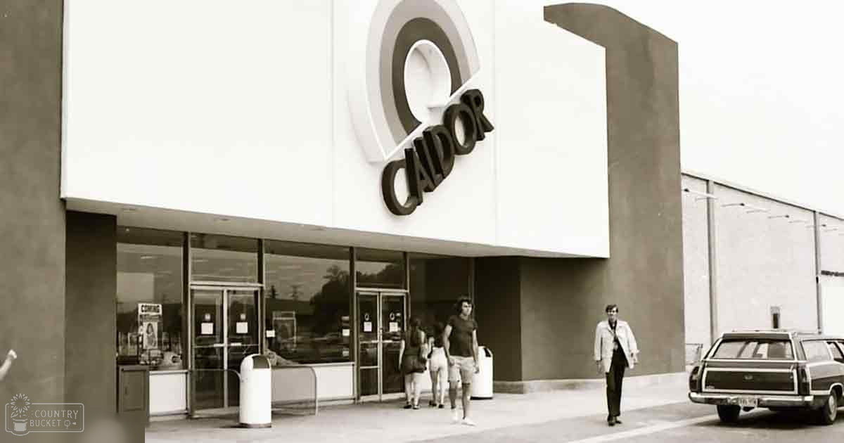 Adult male wearing light jacket over dark shirt and pants; adult female in dark top with shorts and bag slung across body; child in light-colored pants and darker shirt, urban storefront setting with Caldor store front, vintage car parked on right, white cylindrical trash bins near entrance.