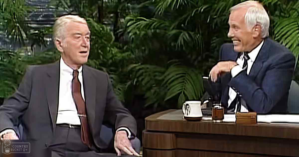 Two older adult men dressed in formal suits with patterned ties, sitting at a round wooden table holding a white mug and dark object, in an indoor talk show setting with lush greenery.