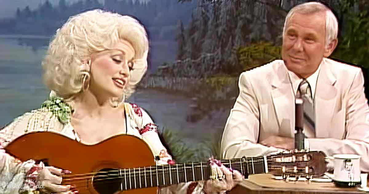 An adult female in a patterned blouse plays guitar with an older male in a light-colored suit jacket, seated against a scenic outdoor backdrop within what appears to be a studio setting.