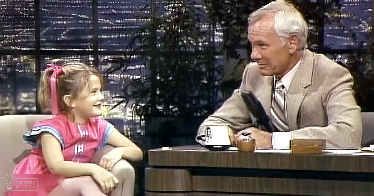 A young female child in a pink dress with white sleeves and blue collar, sitting at an adult male in a beige suit jacket over a patterned tie, both on an indoor talk show set with cityscape background.